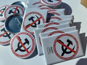 Zestaw białych magnesów prostokątnych i okrągłych przypinek z przekreślonym symbolem sierpa i młota oraz napisem Precz z Komuną, logo Muzeum Zimnej Wojny.